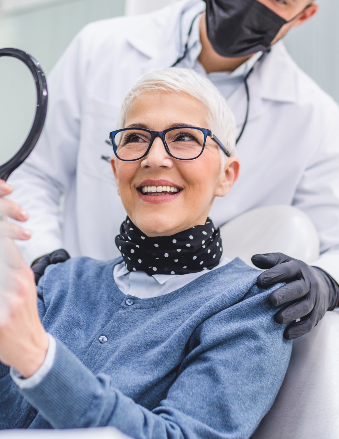 An older woman with white hair and glasses at a dentist office looking at herself in a hand mirror
